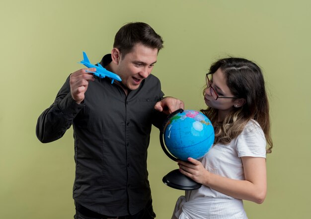 Happy young couple of tourists man and woman holding globe and toy air plane together having fun over light