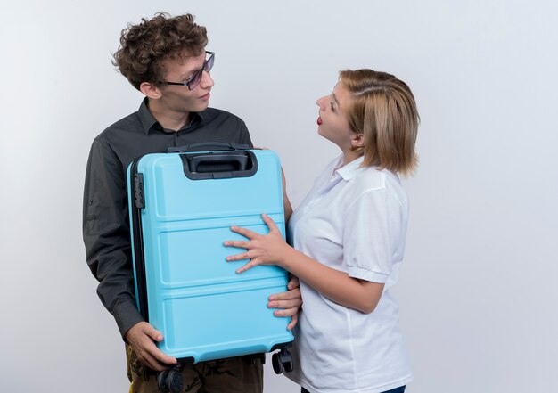 Happy young couple of tourists man and woman holding suitcase looking at each other being surprised standing over white wall