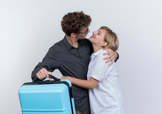 白い壁に抱き合ってお互いを見ているスーツケースを持っている観光客の男性と女性の幸せな若いカップル