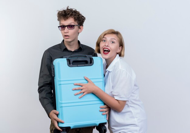 Happy young couple of tourists man and woman holding suitcase looking surprised and amazed standing over white wall