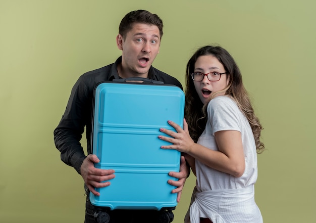 Happy young couple of tourists man and woman holding suitcase looking surprised standing over light wall