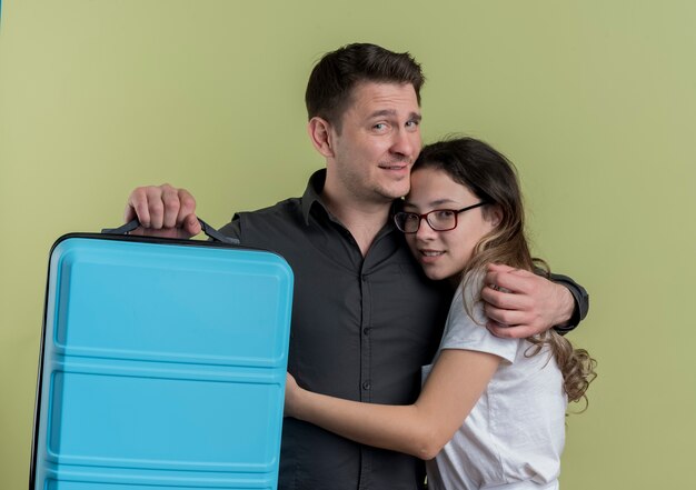 Счастливая молодая пара туристов мужчина и женщина, держащая чемодан, улыбаясь и обнимаясь над светлой стеной