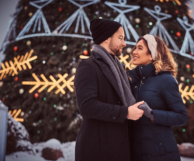 Happy young couple wearing warm clothes hold hands and look at each other, standing near a city Christmas tree, enjoying spending time together.