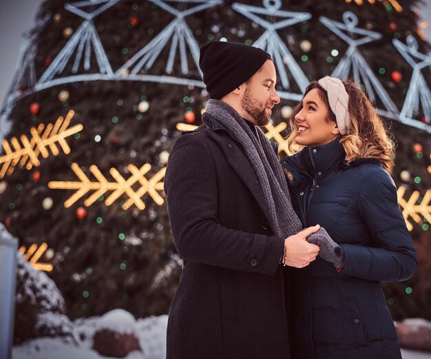暖かい服を着た幸せな若い夫婦が手をつないで見つめ合い、街のクリスマスツリーの近くに立って、一緒に過ごす時間を楽しんでいます。