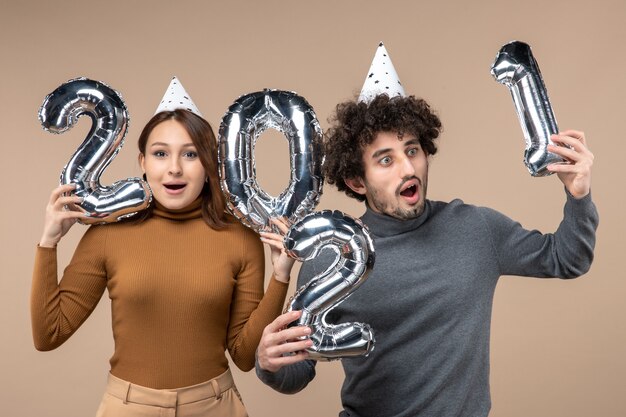 驚いた表情で幸せな若いカップルは、カメラの女の子のショーと灰色の男と新年の帽子のポーズを着用します