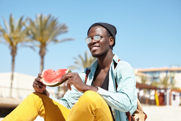 Happy young dark-skinned male traveler wearing stylish clothing sitting on beach and eating watermelon, having relaxed look, enjoying sunny weather during summer vacations in tropical country