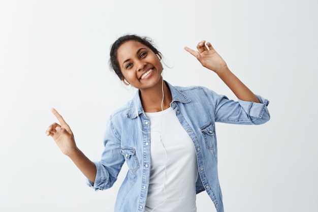 Happy young dark-skinned woman in blue shirt with charming appealing smile, pointing her fingers up having happy look, enjoyind her favourite song and dancing.