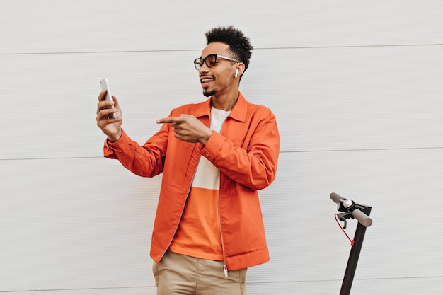 Free photo happy young darkskinned guy in orange jacket colorful tshirt and sunglasses holds phone and talks by video outside