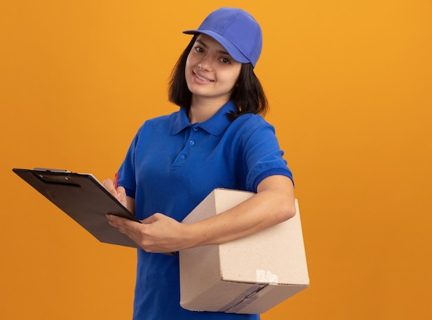 Happy young delivery girl in blue uniform and cap holding cardboard box and clipboard looking  with smile on face standing over orange wall