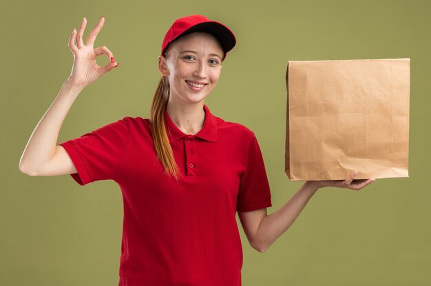 Happy young delivery girl in red uniform and cap holding package   smiling confident doing ok sign