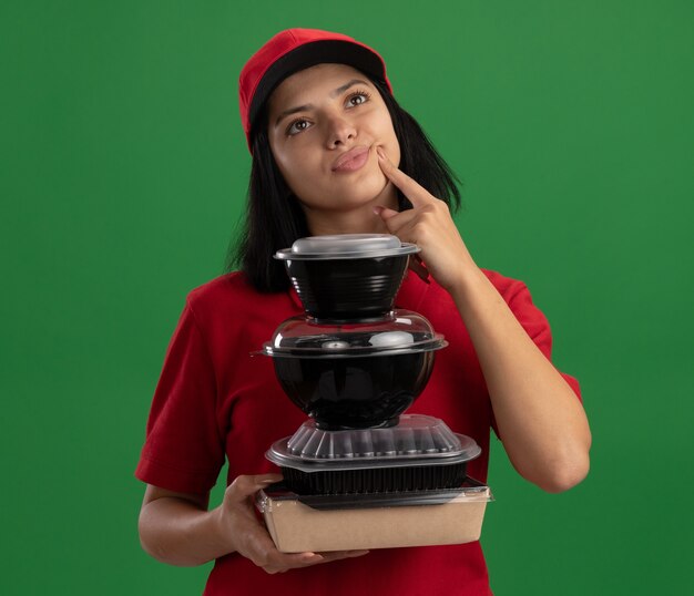 Счастливая молодая доставщица в красной форме и кепке держит стопку продуктовых пакетов, озадаченно глядя в сторону, стоя у зеленой стены