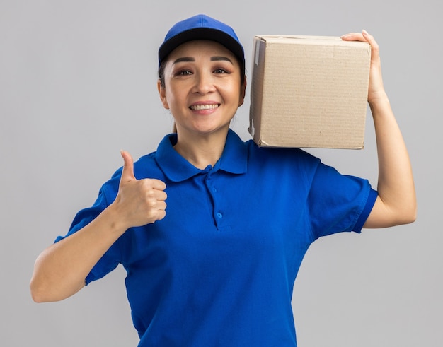 Happy young delivery woman in blue uniform and cap holding cardboard box   with smile on face showing thumbs up  