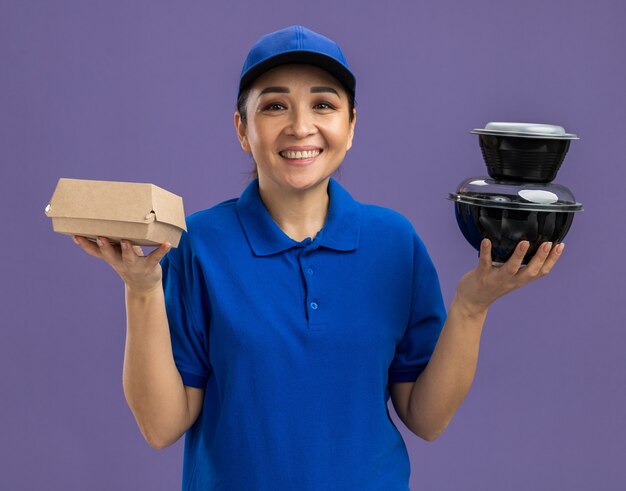 Happy young delivery woman in blue uniform and cap holding food packages  smiling cheerfully standing over purple wall