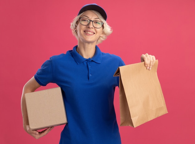 Felice giovane donna delle consegne in uniforme blu e cappuccio che tiene il pacchetto di carta e la scatola di cartone sorridendo allegramente sopra la parete rosa