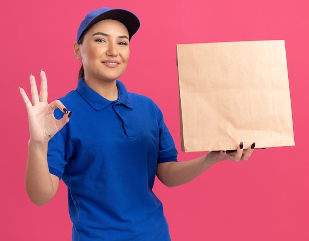 Счастливая молодая женщина-доставщик в синей форме и кепке, держащая бумажный пакет, глядя вперед, весело улыбаясь, показывая знак ОК, стоящий над розовой стеной