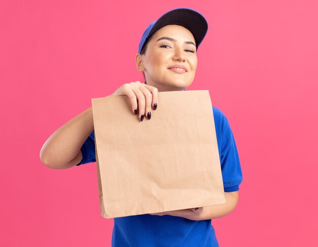 Happy young delivery woman in blue uniform and cap holding paper package looking at front smiling cheerfully standing over pink wall