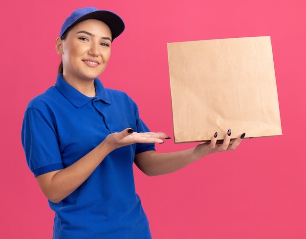 Happy young delivery woman in blue uniform and cap holding paper package presenting with arms looking at front smiling cheerfully standing over pink wall