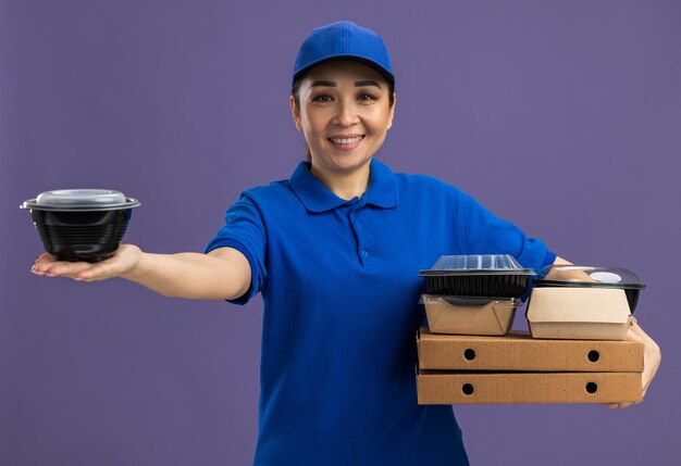 青い制服を着た幸せな若い配達女性と、ピザの箱と食品のパッケージを持った帽子が、紫色の壁の上に立って陽気に笑っている