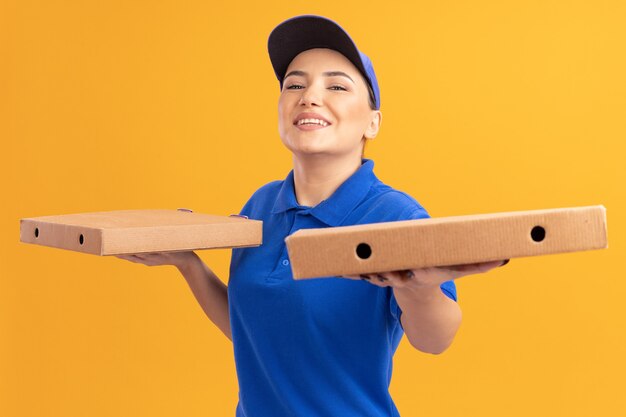 Happy young delivery woman in blue uniform and cap holding pizza boxes looking at front smiling cheerfully standing over orange wall