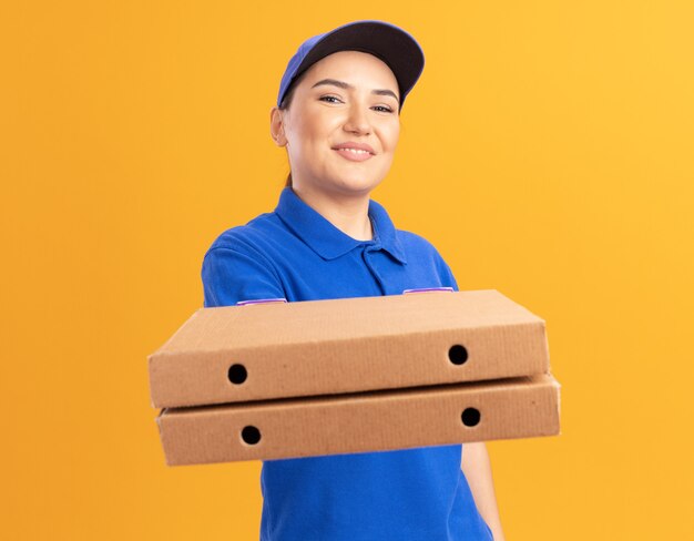 Счастливая молодая женщина-доставщик в синей форме и кепке, держащая коробки для пиццы, глядя вперед, уверенно улыбаясь, стоя над оранжевой стеной