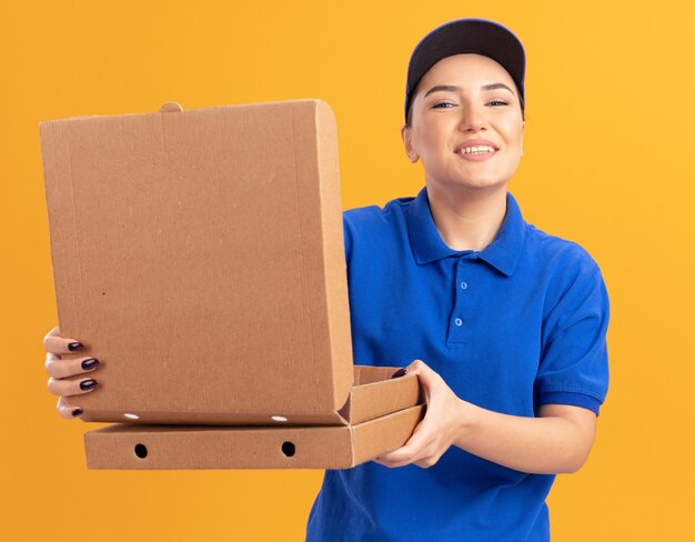 행복 한 젊은 배달 여자 파란색 유니폼과 모자를 들고 피자 상자 열기 상자 앞에 오렌지 벽 위에 유쾌하게 서 웃 고보고