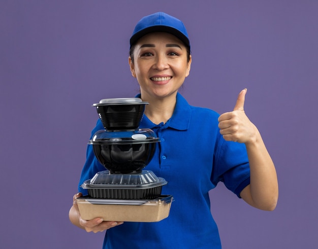 Happy young delivery woman in blue uniform and cap holding stack of food packages  smiling cheerfully showing thumbs up standing over purple wall