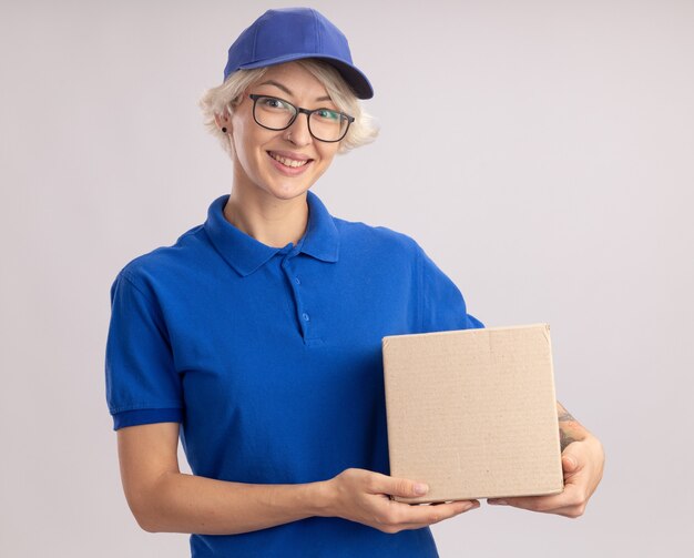 Happy young delivery woman in blue uniform and cap wearing glasses holding cardboard box  smiling cheerfully standing over white wall