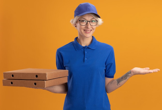 파란색 유니폼과 모자 오렌지 벽 위에 유쾌하게 웃는 피자 상자를 들고 안경을 쓰고 행복 젊은 배달 여자