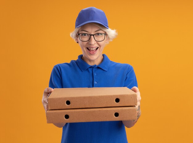 青い制服を着た幸せな若い配達の女性とオレンジ色の壁に元気に笑ってピザボックスを保持している眼鏡をかけて