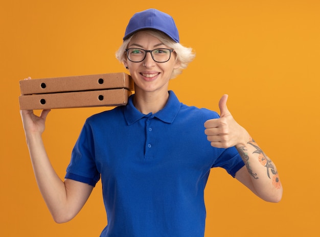 Felice giovane donna delle consegne in uniforme blu e berretto con gli occhiali in possesso di scatole per pizza sorridente che mostra i pollici sopra la parete arancione