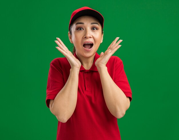 Happy young delivery woman in red uniform and cap  amazed and surprised standing over green wall
