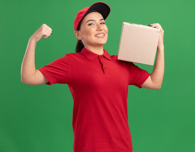 Счастливая молодая женщина-доставщик в красной форме и кепке держит картонную коробку, глядя вперед, весело улыбаясь, поднимая кулак, как победитель, стоящий над зеленой стеной