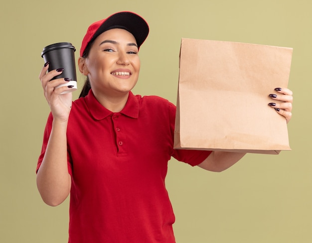 녹색 벽 위에 서있는 얼굴에 미소로 정면을보고 빨간 유니폼과 모자를 들고 종이 패키지와 커피 컵에 행복 젊은 배달 여자