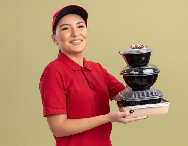 Happy young delivery woman in red uniform and cap holding stack of food packages looking at front smiling cheerfully standing over green wall