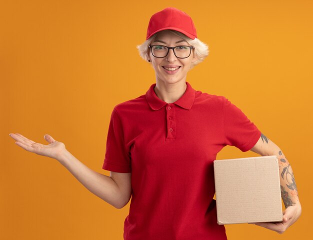 オレンジ色の壁の上に立っている彼女の手の腕でコピースペースを提示して笑顔の段ボール箱を保持している眼鏡と赤い制服を着た幸せな若い配達の女性