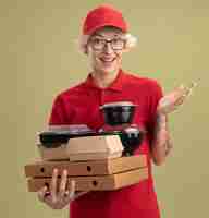 Free photo happy young delivery woman in red uniform and cap wearing glasses holding pizza boxes and food packages smiling  standing over green wall