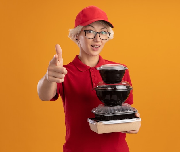 Счастливая молодая женщина-доставщик в красной форме и кепке в очках держит стопку продуктовых пакетов, указывая указательным пальцем, уверенно улыбаясь, стоя над оранжевой стеной