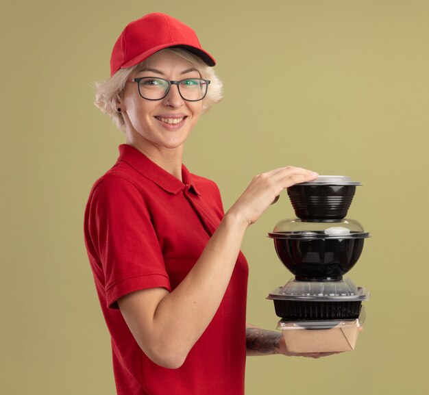 Happy young delivery woman in red uniform and cap wearing glasses holding stack of food packages  smiling confident standing over green wall