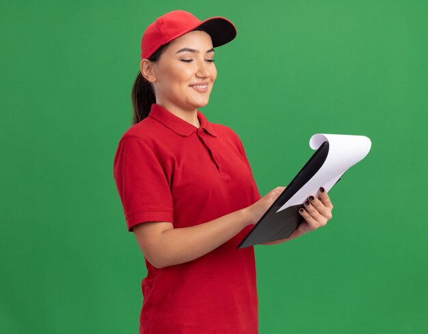 Happy young delivery woman in red uniform and cap with clipboard looking at it with smile on face standing over green wall