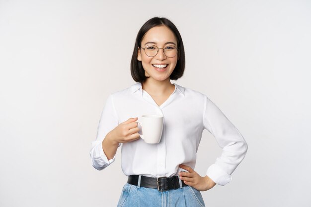 Счастливая молодая энергичная азиатская женщина улыбается, пьет, держа чашку кофе, стоя уверенно на белом фоне