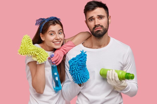 Happy young European woman and dissatisfied tired man hold detergents and rags, clean up room, dressed in white casual clothing, pose over pink wall. Housekeeping and cleanliness concept