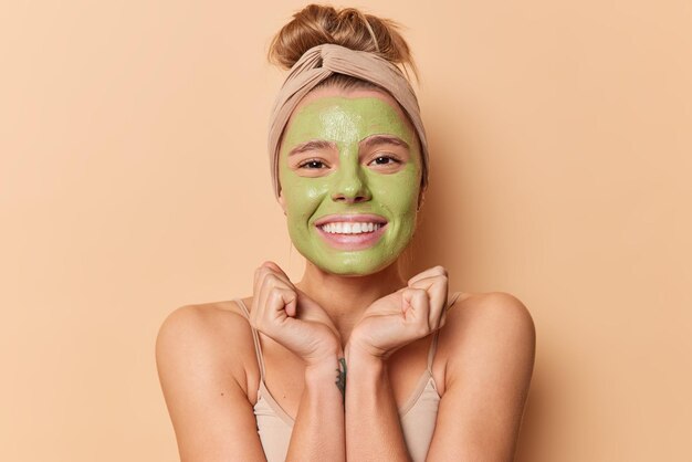 행복한 젊은 유럽 여성은 턱 미소 아래 손을 유지하고 녹색 영양 마스크를 기꺼이 적용하고 머리띠는 베이지색 배경 위에 격리된 피부 치료를 위해 미용 절차를 받습니다.