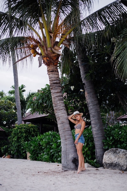 Felice giovane donna europea in pantaloncini di jeans bikini top leopardo e cappello bianco classico sulla spiaggia esotica tropicale sorridente in posa divertendosi ragazza giocosa sul concetto di viaggio di vacanza