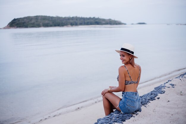 Happy young european woman in leopard top bikini jean shorts and classic white hat on tropical exotic beach smiling posing having fun Playful girl on vacation travel concept