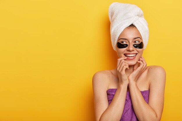 happy young European woman touches skin gently, looks aside, applies beauty undereye patches, wrapped in towel, looks on left side