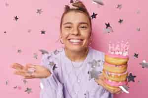 Free photo happy young european woman with combed hair toothy smile holds pile of sweet doughnuts with burning candles poses against pink background flying stars around celebrates birthday waits for guests