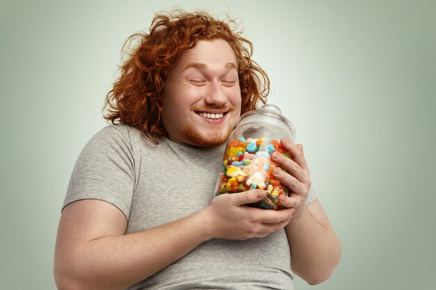 嬉しそうに笑って、目を閉じて幸せな若いデブ肥満男がグッズのガラス瓶で喜び