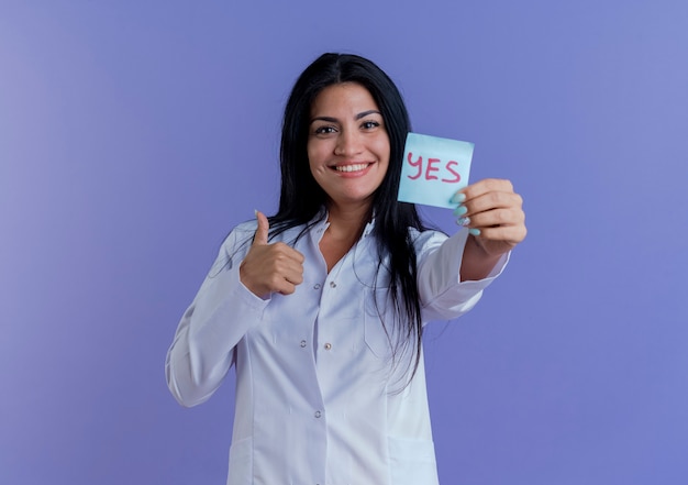 Happy young female doctor wearing medical robe showing yes note, looking showing thumb up