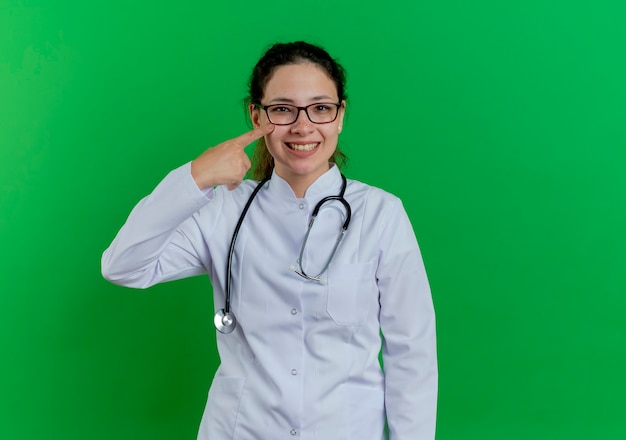 의료 가운과 청진 기 및 복사 공간이 녹색 벽에 고립 된 뺨에 손가락을 가리키는 안경을 착용하는 행복 한 젊은 여성 의사