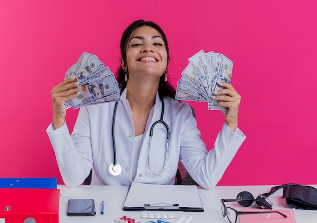 Foto gratuita felice giovane medico femminile che indossa abito medico e stetoscopio seduto alla scrivania con strumenti medici che tengono soldi isolati sulla parete rosa